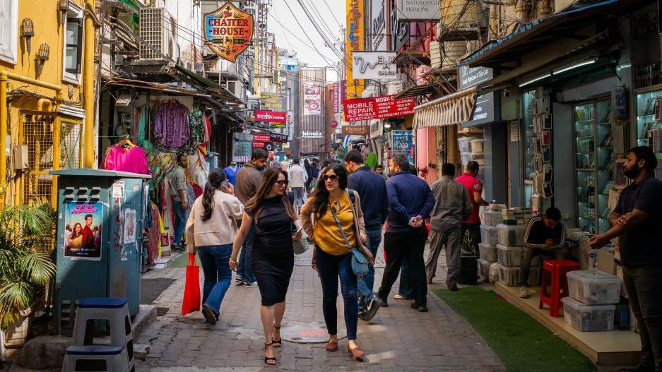 The image shows two women wearing sun glasses pass stores at a bustling market in New Delhi, India
