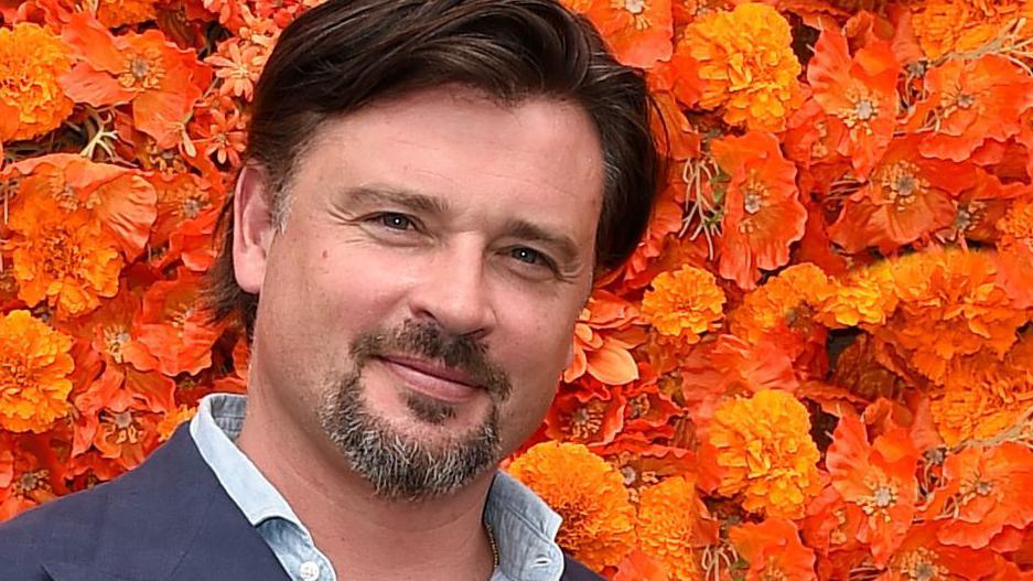 Actor Tom Welling standing in front of a wall of orange flowers at a press junket. He has a brown and grey goatee beard, dark hair and wears a navy jacket and a chambray shirt.