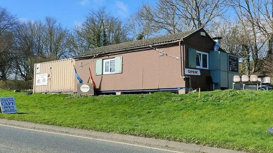 A cabin at the side of the road which is a cafe, it sits on a grassy bank and has chairs outside and a sign which says 'cafe open'