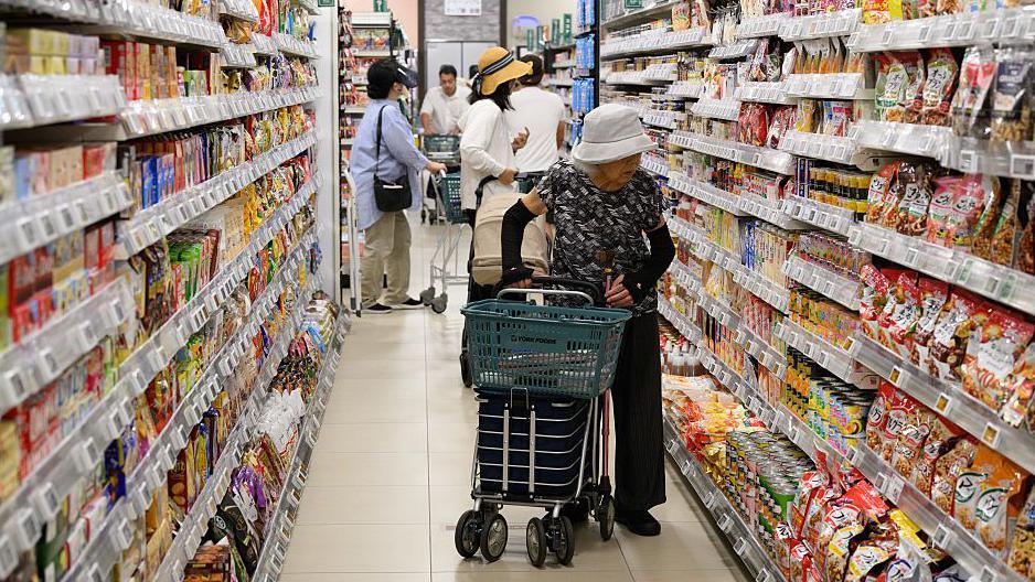An elderly woman wearing a white hat and dark clothing browses the crowded shelves at a Japanese supermarket, as she pushes a shopping trolley.