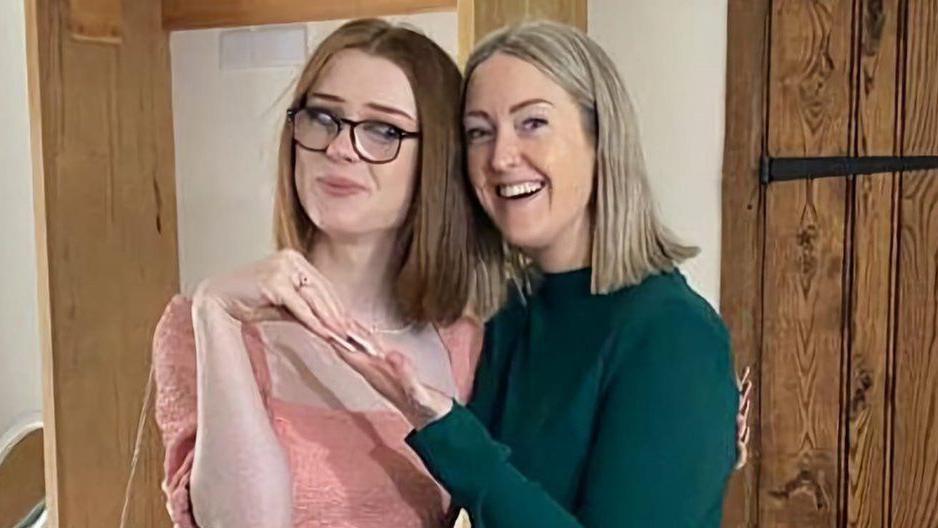 Brianna Ghey, who has long straight dark reddish brown hair and glasses, and is wearing a pink top, poses for a picture next to Esther Ghey who has straight blonde hair and is wearing a dark green jumper. 