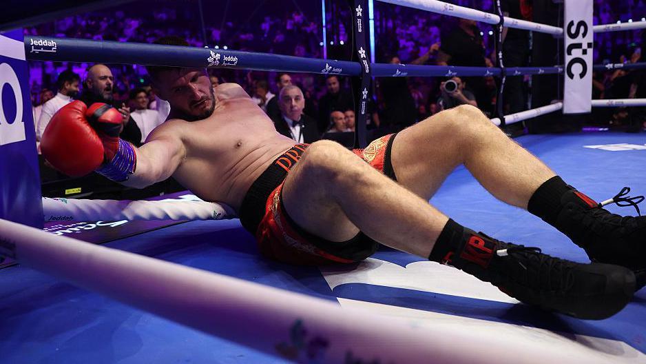 Kristian Prenga is grounded, with his back against a rope and his head partway outside the boxing ring