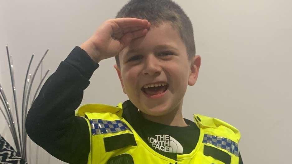 Archie give a salute with his right hand held up to his forehead while wearing a black top and toy police vest. The vest is bright yellow and had blue and white checked badges across the shoulders