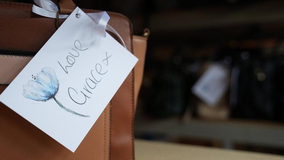 The corner of a brown leather handbag with a white tag attached. The tag has a flower on it and reads "Love Grace x".