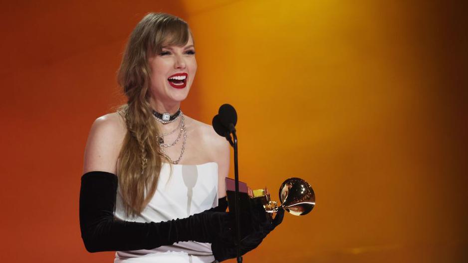 Taylor Swift wearing a white dress with black gloves holds a Grammy Award at the 2024 ceremony.