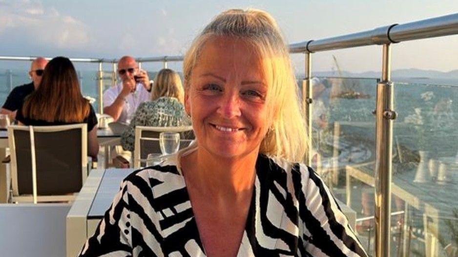 Anita Rose is wearing a black and white zebra print shirt, with her long blonde hair in a ponytail. She is sat at a table at a rooftop bar. She's smiling and looking directly at the camera.