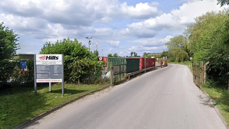 A view looking down the road into the Hills Lower Compton Waste site. A sign for it to the left, with shipping containers behind a fence and green trees down the right hand side.