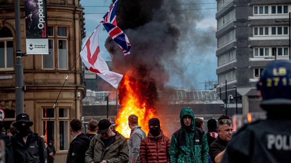 A group of men stand in front of a fire during the riots in Sunderland. A union flag and a Cross of St George flies above the scene as police officers look on.