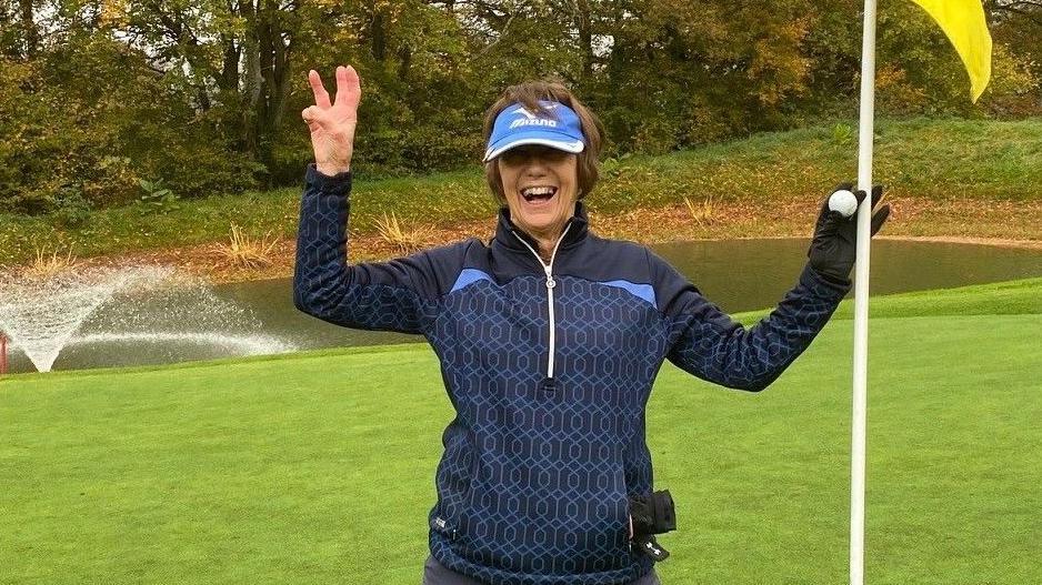 Lyn Parry holds up three fingers and a flag to celebrate her hole-in-one treble success