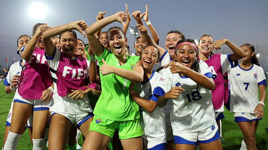 Samoan players in white and blue uniforms celebrate on the field. They gesture with raised arms and point upward after a match, with stadium lights and flags in the background.