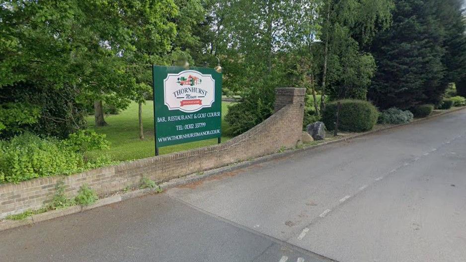 A sign at the entrance to the car park of Thornhurst Manor Country Park. It is next to a small brick wall and surrounded by trees.