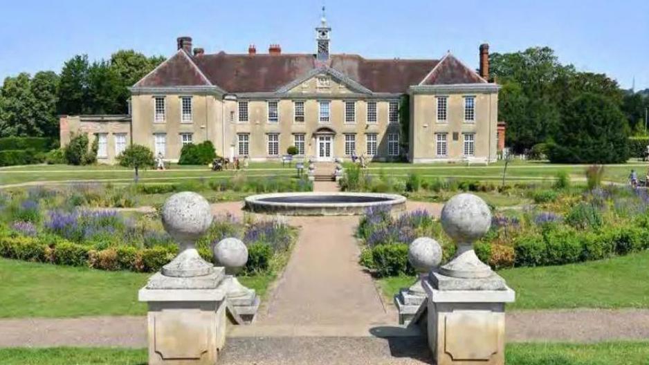 A light brown two-floor building in grass grounds. There is a water feature in the centre of the image.