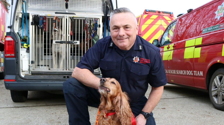 Graham Currie kneels behind service dog Jarvis. Mr Currie is wearing a navy fire service uniform while Jarvis is a chestnut coloured dog with a red and orange coloured collar and lead on him. Fire service vehicles are parked behind them.