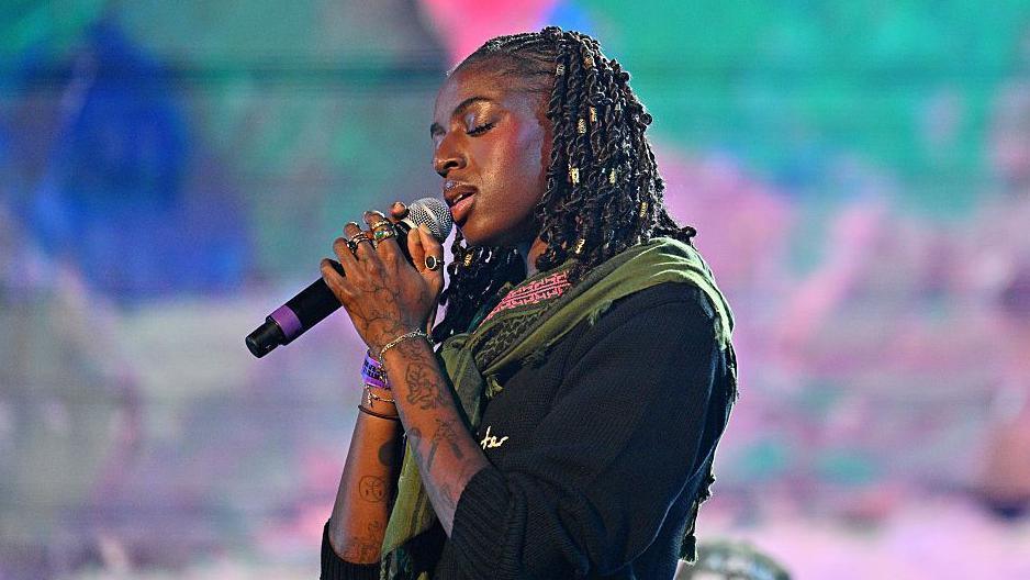 Cat Burns sings on-stage. Her eyes are closed as she clasps the microphone.