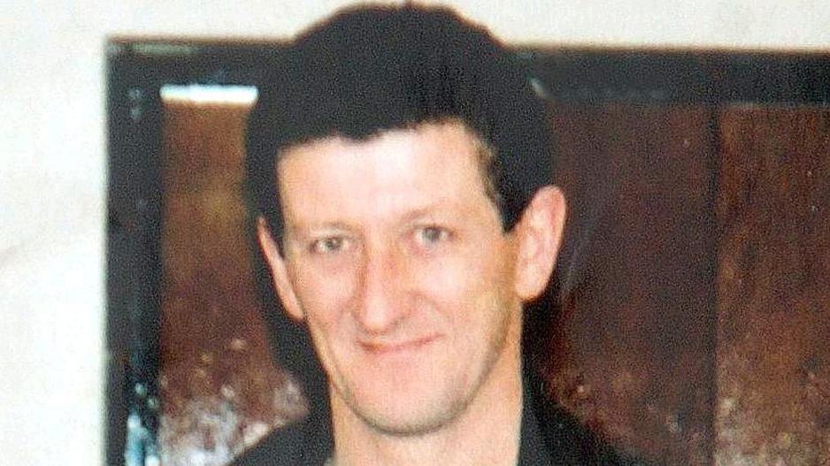 A smiling Jim McFadden stands against a brown background. He has short black hair and is wearing a black jacket.