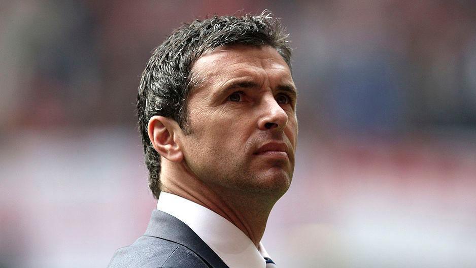 Gary Speed looks upwards during a 2011 match between Wales and England in the stadium. He is wearing a dark suit and tie.