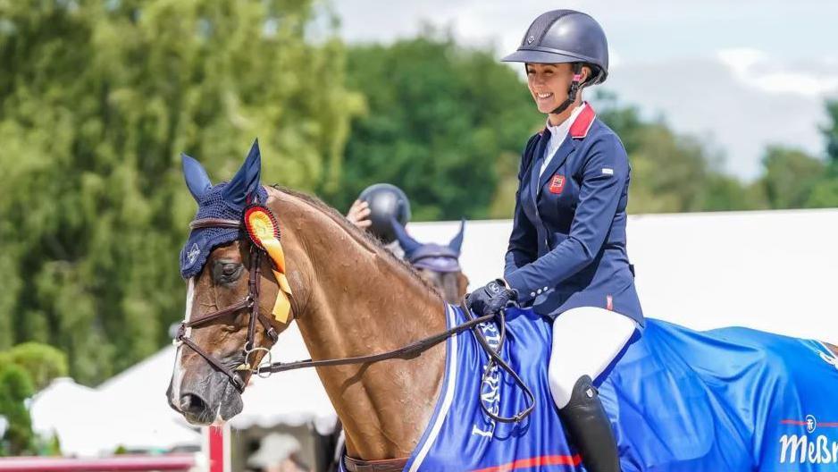 Yasmin Ingham on horseback holding the reins. She is wearing a navy blue helmet, a navy jacket, white jodphurs and black boots.