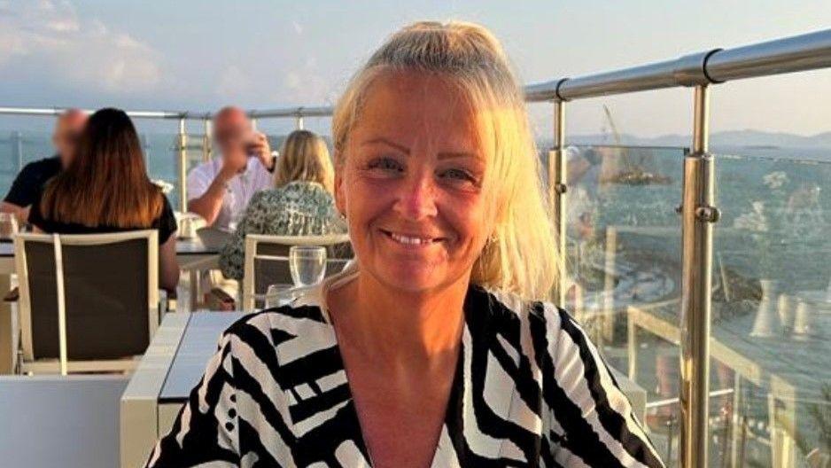 A woman smiling directly into the camera. She is sitting at a table in a restaurant on a terrace. Other diners are in the background.