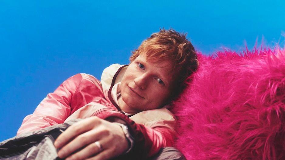 Ed Sheeran reclines on a fluffy pink chair, with his head turned to the camera. He is smiling and has ginger hair.