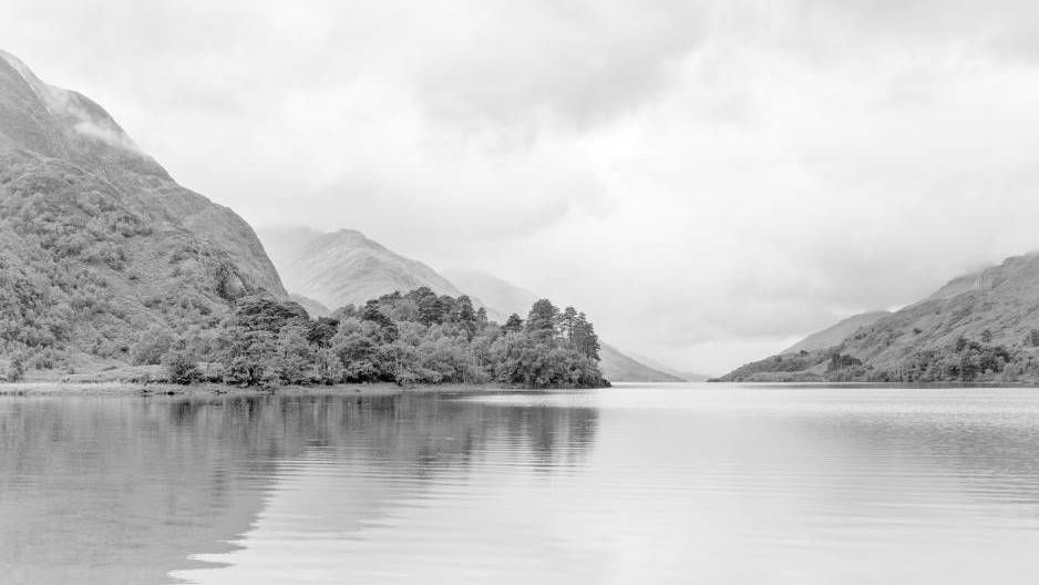 A black and white image of a loch. There are trees in the distance and clouds in the sky.