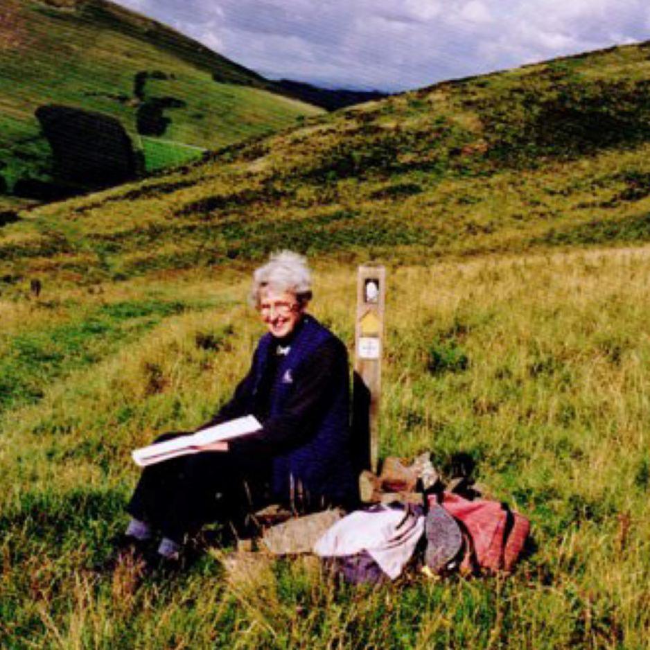 Audrey Henshall is sitting on rocks next to a waymarker for a path. She has a map on her lap. She is surrounded by a landscape of green hills.