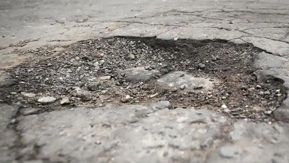 A pothole is seen in the close-up image. It has gravel in the dip and patchy bits of concrete.