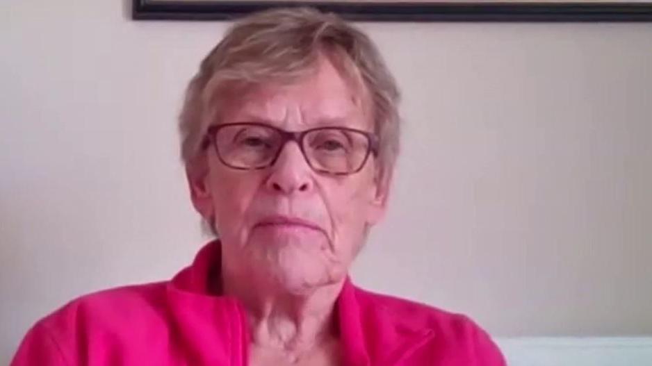 A woman sitting on a chair wearing a pink fleece. She has mousey-coloured hair and is wearing glasses and looking directly at the camera. There is a radiator in the background.