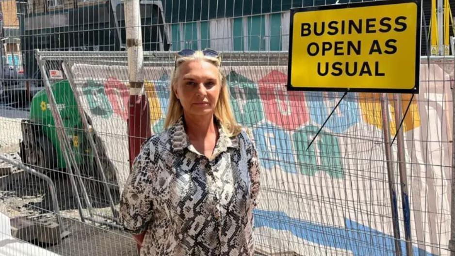Julie Goodwin has long blonde hair and has sunglasses on top of her head. She is wearing a grey and white patterned shirt and is standing in from of metal roadworks fencing. There is a yellow sign with black writing which reads Business open as usual.