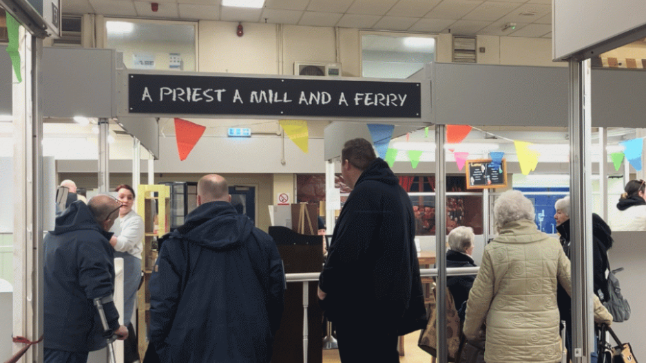 People queuing at the A Priest a Mill and a Ferry cafe. There are five people in the queue, each with their backs to the camera. There is multi-coloured bunting up in the cafe. 