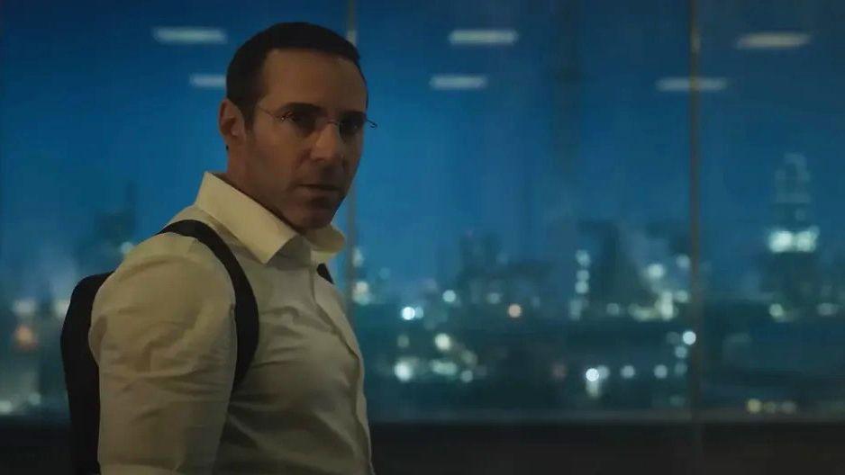 A still from the film showing a man with dark hair, glasses wearing a white shirt and backpack stood in front of windows behind which is an image of the refinery at night