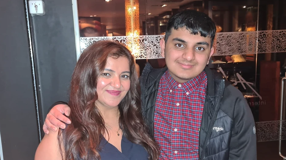 Pooja and Ronan are standing outside a restaurant. Both are smiling with Rona's arm around his mother. She has long dark hair and a sleeveless top on and Rona has a checked shirt on and a black jacket.