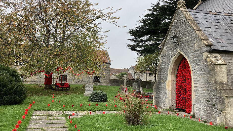 A church with knitted and crocheted poppies lining the path and covering the large wooden door. There are also poppies in a tree and covering a bench in the graveyard. 