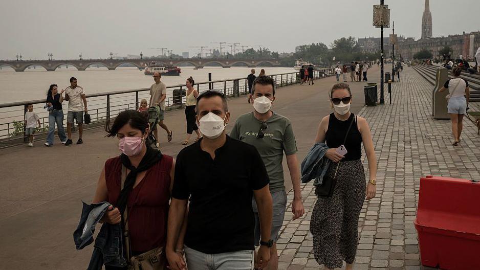 People walking around with face masks on in Bordeaux, along a riverside cloaked in deep grey smoke