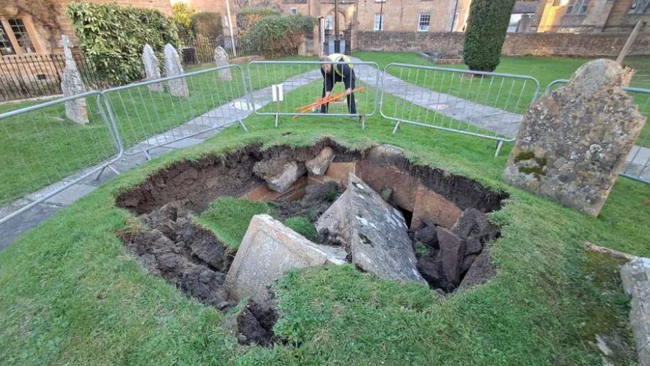 Image shows a hole at the edge of a graveyard with graves that have fallen inside. A man in a florescent vest can be seen near the back of the collapsed tomb, looking in. 