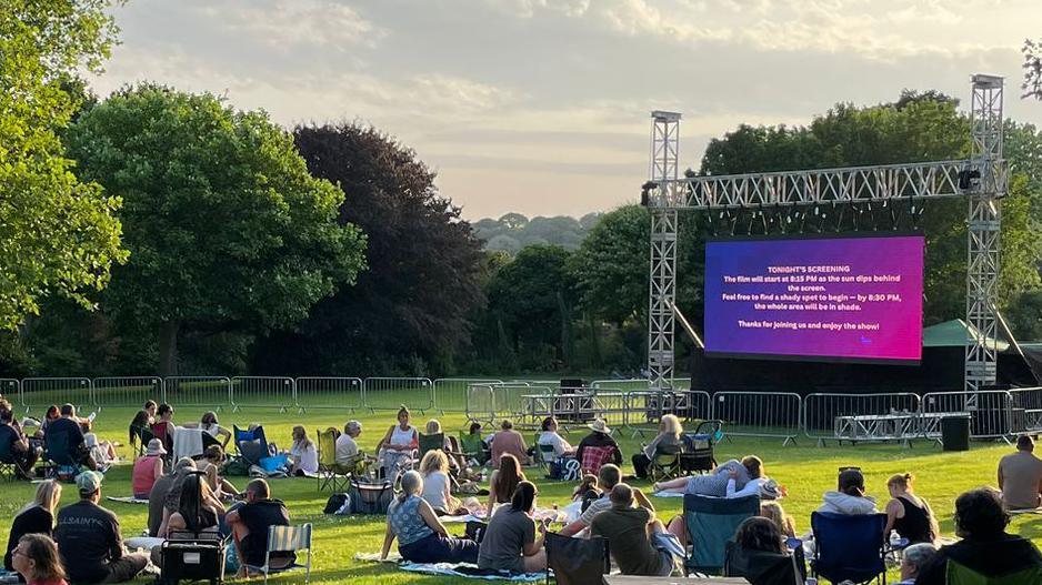 People sat in deck chairs and on picnic blankets in front of a big screen on a grassy area of a park. There are trees around the park.