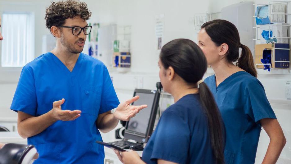 One male and two female nurses in blue scrubs stand in a hospital room.