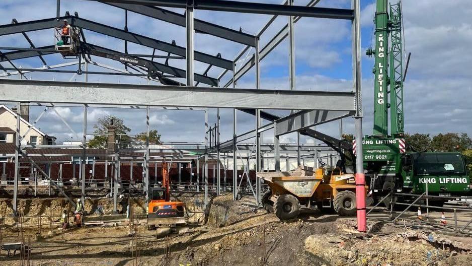 Image shows a large steel structure with JCBs and a crane underneath in a dirt area. This is the leisure facility structure. 