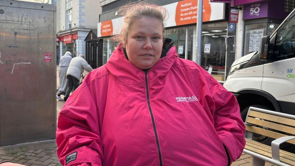 A woman with light brown hair tied in a ponytail, wearing a pink coat.She is standing on the street, with shops pictured behind her.