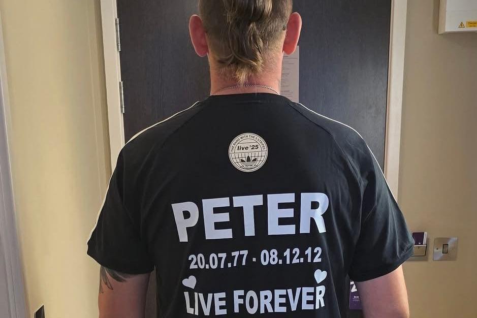 Man standing with his back to the camera with a black T-shirt featuring the words: "Peter 20.07.77 - 08.12.12 Live Forever" printed in white on the back.