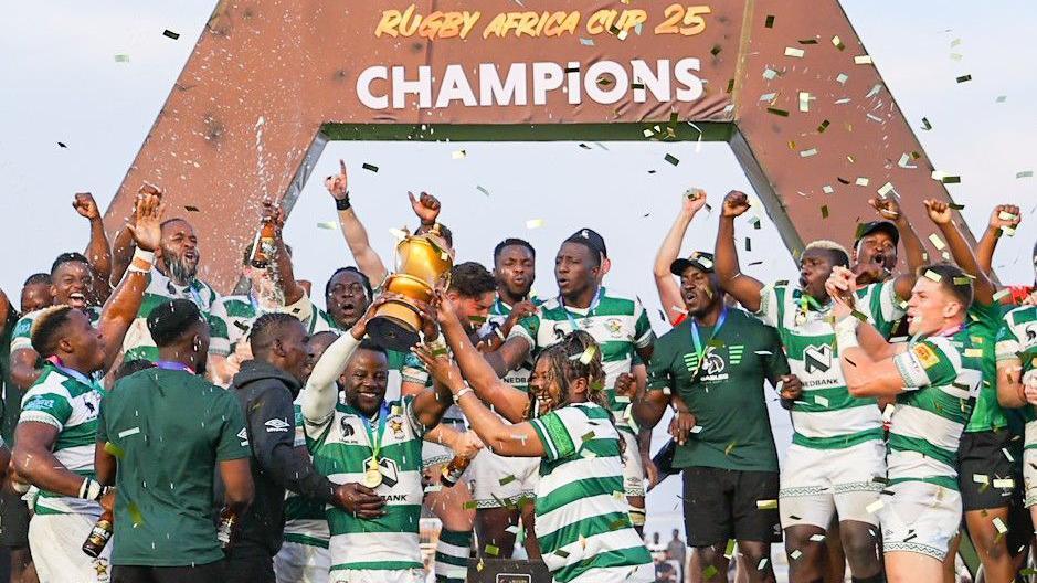 Zimbabwe's rugby players dressed in green and white lift a golden trophy and celebrate in front of a sign that says Rugby Africa Cup 25 champions
