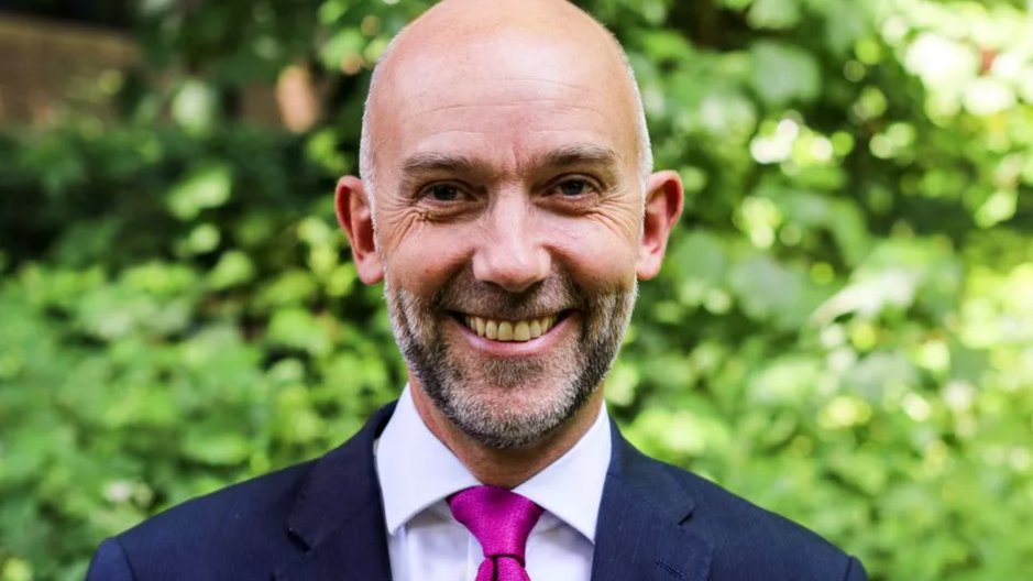 Adrian Crompton, looking straight at the camera and smiling. He has a neatly trimmed short beard, bald head and is wearing a magenta tie, white shirt and dark blue suit jacket
