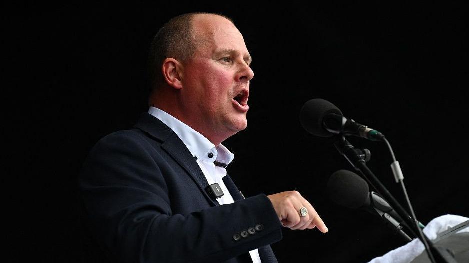 TUC general secretary Paul Nowak addresses the Durham Miners' Gala, dressed in a dark suit with an open neck shirt. He is making a point with his finger and speaking into two microphones