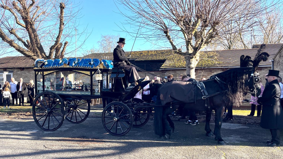 A stationary funeral carriage drawn by two black horses is attended by two men in black top hats, while groups of people can be seen in the background.
