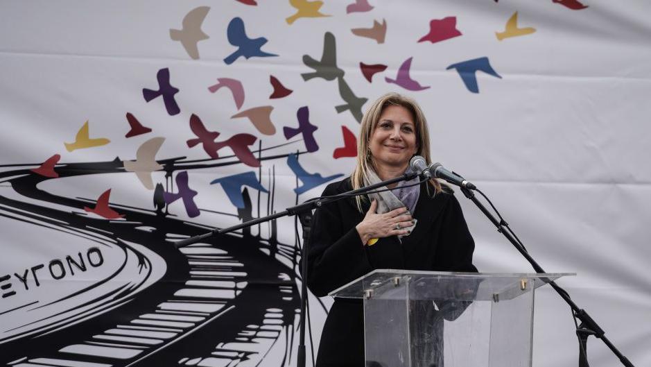The mother of a victim stands with her hand against her chest against a white backdrop decorated with flying birds