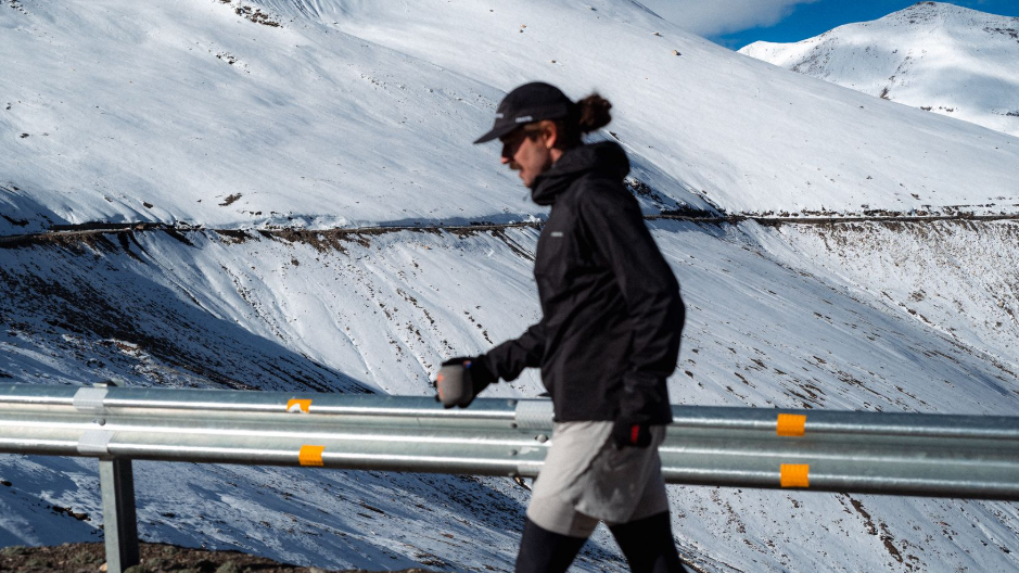 Landscape shot of Jack in his running gear on a snow-covered mountain pass. He is wearing gloves, a hat and holding a coffee mug in his right hand