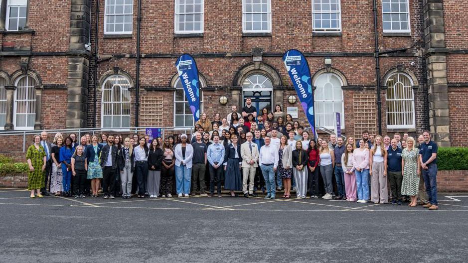 A crowd of staff and students standing outside Pears Cumbria School of Medicine. The school is based at the University of Cumbria's Fusehill Street campus in Carlisle. It is a two-storey redbrick building.