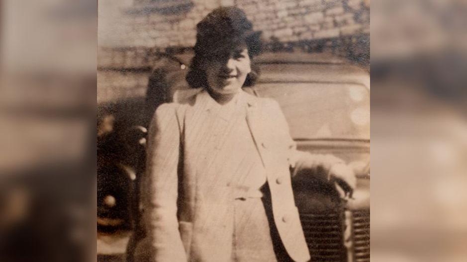 A black and white photo of Joyce in her youth leaning up against the hood of a car wearing a dark hat and blazer