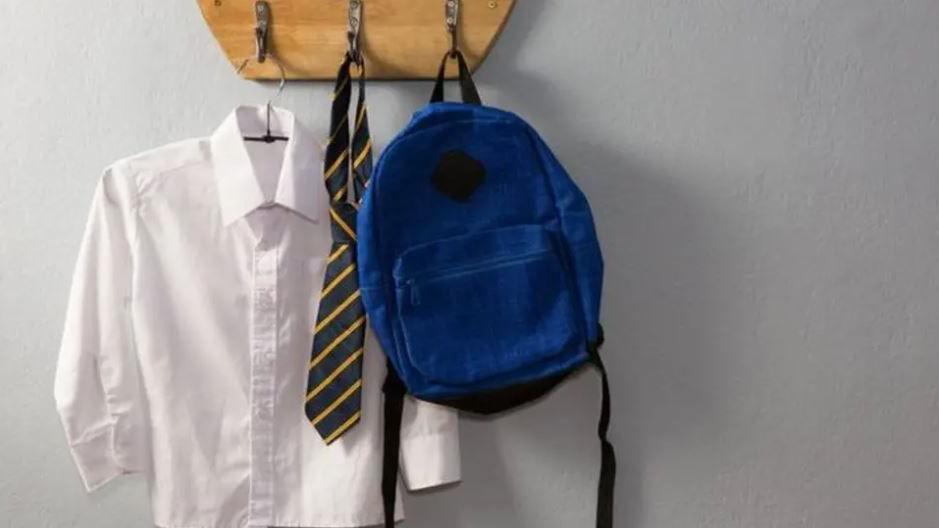 A white shirt, tie with yellow stripes and blue rucksack hanging from metal pegs on a wooden placard on a wall.