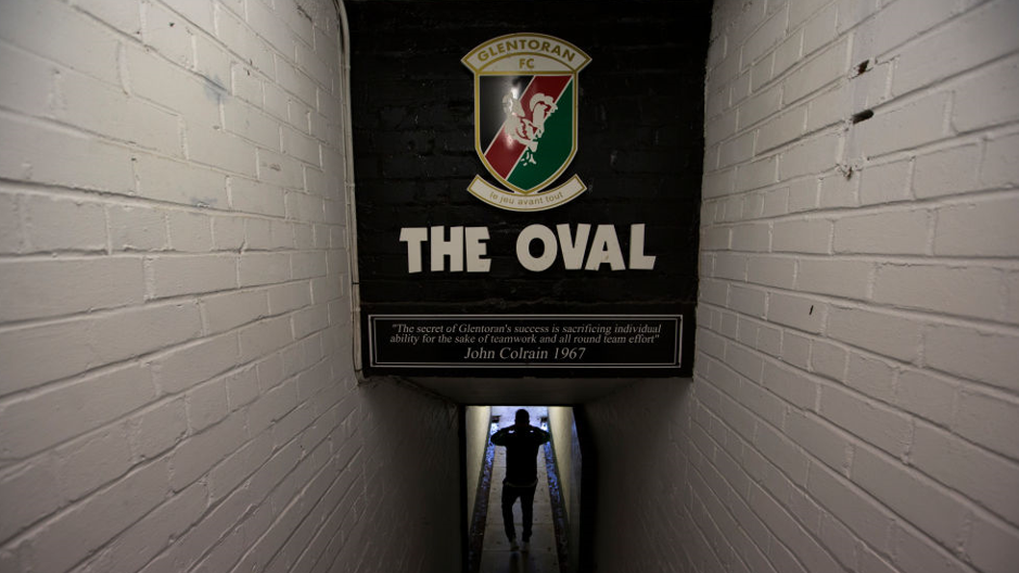 Man walks under an image saying The Oval, with a quote from former manager John Colrain underneath. There are two white walls on either side.