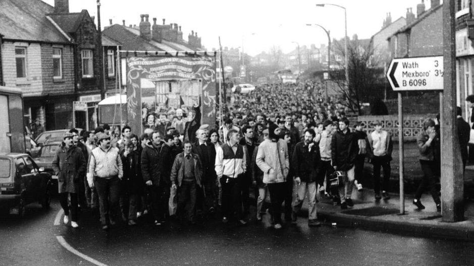 A black and white picture showing a large group of men walking along the street. There is a banner being held up in the air by some of the men. Houses line the road and there is a road sign showing the distance to Wath and Mexborough.